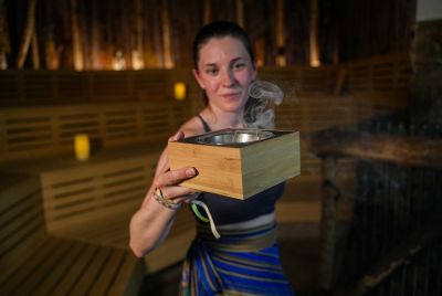 Eine lächelnde Saunameisterin hält in einer Holzsauna eine Holzbox mit einer dampfenden Schale für ein aromatisches Ritual in den Händen.