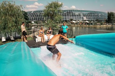 Ein Mann surft auf einer künstlichen Surfwelle mit der Therme Euskirchen im Hintergrund