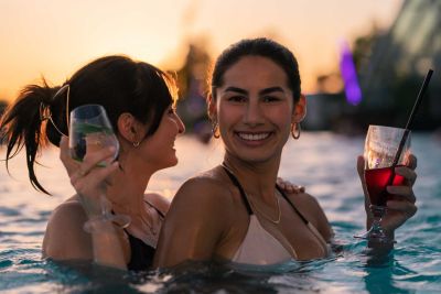 Zwei Frauen tanzen und lachen im Pool mit Cocktails in der Hand