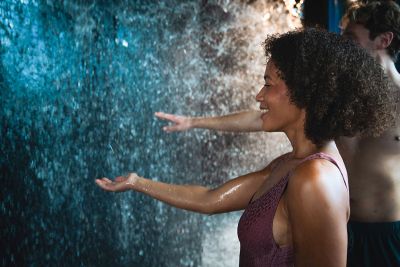 A smiling woman relaxes, stretching her hands out towards a gentle rain of water droplets.