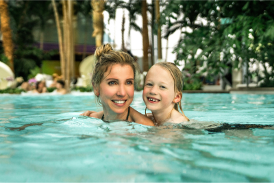 Eine lächelnde blonde Frau und ein junges Mädchen schwimmen eng nebeneinander im hellblauen Wasser eines Thermalbades, umgeben von tropischen Pflanzen im Hintergrund.