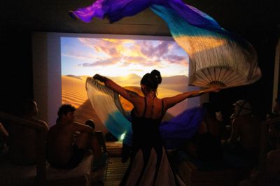 Eine Aufgusskünstlerin von hinten bei einer Show-Performance mit wehenden, farbenfrohen Fächern vor einer beleuchteten Wüstenkulisse im Saunaraum.