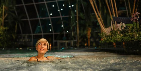 Eine lächelnde Frau mit kurzen blonden Haaren liegt im Wasser und blickt nach oben. Im Hintergrund erstreckt sich eine große, gewölbte Glasdachkonstruktion, an der kleine Lichter im Dunkeln leuchten, umgeben von Pflanzen.