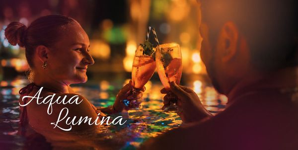 Eine lächelnde Frau blickt im stimmungsvoll beleuchteten Wasser zu ihrem Gegenüber und stößt mit einem Cocktailglas an. Im Hintergrund reflektieren goldene und grüne Lichter auf der Wasseroberfläche. Unten links steht der geschwungene, weiße Schriftzug „Aqua Lumina“.