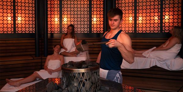 Ein Saunameister führt in einer atmosphärisch beleuchteten Sauna einen Aufguss durch, während Gäste im Hintergrund entspannen.