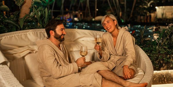 Mann und Frau sitzen in gemütlichen Bademänteln in einer hellen Relaxmuschel und stoßen lächelnd mit Getränken an.